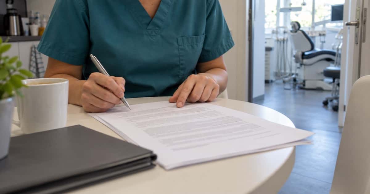 Hände einer ZFA im Kasack mit Stift über einem ausgedruckten Arbeitsvertrag im Pausenraum der Praxis